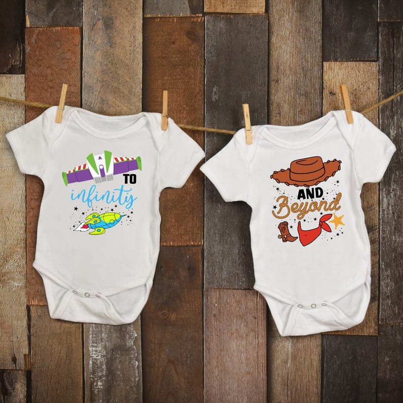 May include: Two white baby onesies with cartoon graphics. One says "To Infinity" with a spaceship design. The other says "And Beyond" with a cowboy hat and boot design. Both are hanging on a wooden background.