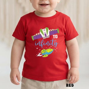May include: A red t-shirt featuring a colorful graphic of a rocket and the phrase "To infinity" in blue script. The design includes a rocket ship and a stylized space vehicle. The word "RED" is printed at the bottom.