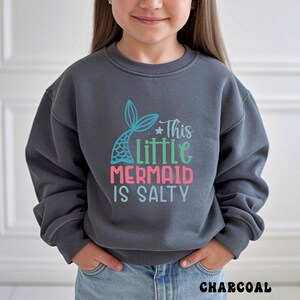 Puede incluir: Una sudadera gris carb&oacute;n con el texto "This Little Mermaid Is Salty" en turquesa, verde y rosa. Un gr&aacute;fico de cola de sirena est&aacute; a la izquierda. La palabra "CHARCOAL" est&aacute; en la parte inferior. La sudadera la lleva un ni&ntilde;o.