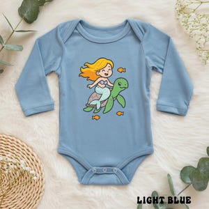 Puede incluir: Body de beb&eacute; azul claro con mangas largas. Presenta una sirena de dibujos animados montada en una tortuga verde, rodeada de peces naranjas. El texto "LIGHT BLUE" est&aacute; impreso en la parte inferior.