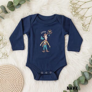 May include: Navy blue baby bodysuit with long sleeves. The front features a cartoon illustration of Woody from Toy Story, with the word "Woody" above the character. The design includes a sun and smoke effect. The bodysuit has three snap closures.