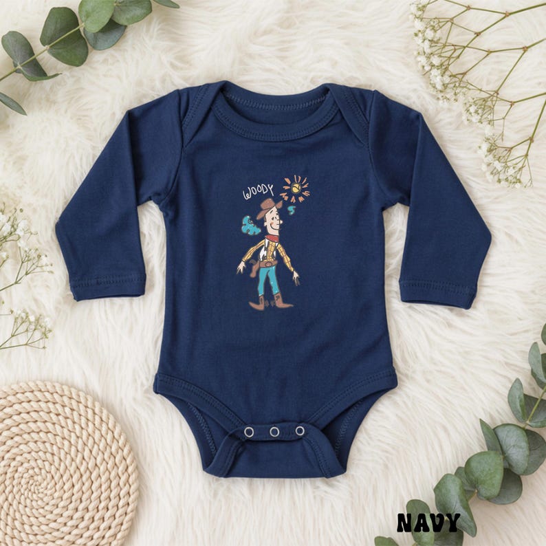 May include: Navy blue baby bodysuit with long sleeves. The front features a cartoon illustration of Woody from Toy Story, with the word "Woody" above the character. The design includes a sun and smoke effect. The bodysuit has three snap closures.