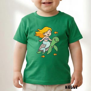 Puede incluir: Camiseta verde con una sirena de dibujos animados montada en una tortuga verde, rodeada de peque&ntilde;os peces naranjas. La sirena tiene pelo rubio y una cola azul. La palabra "KELLY" est&aacute; impresa en la parte inferior.