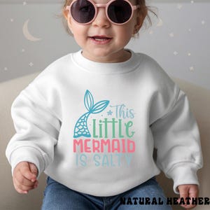 Puede incluir: Sudadera blanca con un dise&ntilde;o de cola de sirena y el texto "This Little Mermaid Is Salty" en azul, verde y rosa. La sudadera est&aacute; hecha de un material suave.