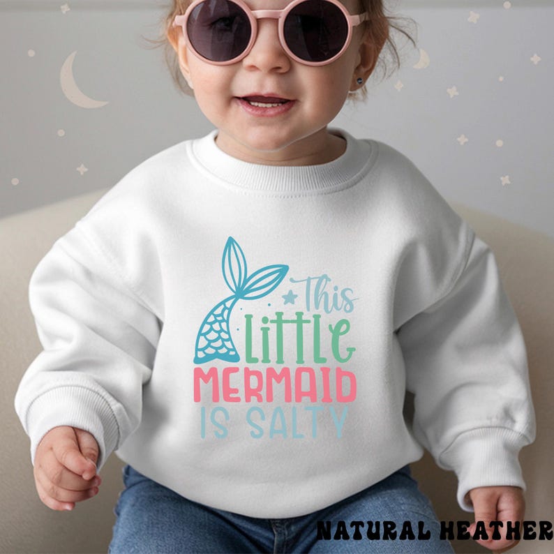 Puede incluir: Sudadera blanca con un dise&ntilde;o de cola de sirena y el texto "This Little Mermaid Is Salty" en azul, verde y rosa. La sudadera est&aacute; hecha de un material suave.