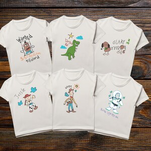 May include: Six white t-shirts with cartoon character designs. One shirt has "Wanted" and "$50 Billion Reward" text. Other shirts feature a dinosaur, a dog, a cowgirl named Jessie, Woody, and Buzz Lightyear. The shirts are on a wooden background.