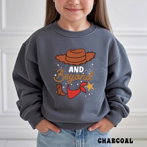 May include: Charcoal gray sweatshirt with a cowboy-themed graphic. The design includes a brown cowboy hat, the words "AND Beyond" in a decorative font, a cowboy boot, a red bandana, and a gold star. The word "CHARCOAL" is printed at the bottom.