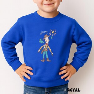 May include: Royal blue sweatshirt featuring a cartoon illustration of Woody from Toy Story. The design includes Woody in his cowboy attire, with the word "WOODY" above him. The sun and a few clouds are also depicted.