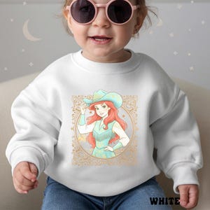 K&ouml;nnte beinhalten: Wei&szlig;es Sweatshirt mit einer Cartoon-Illustration einer Frau mit Cowboyhut und t&uuml;rkisfarbener Weste. Die Illustration ist in einem goldenen, verzierten Design gerahmt. Das Wort "WHITE" ist unten aufgedruckt.