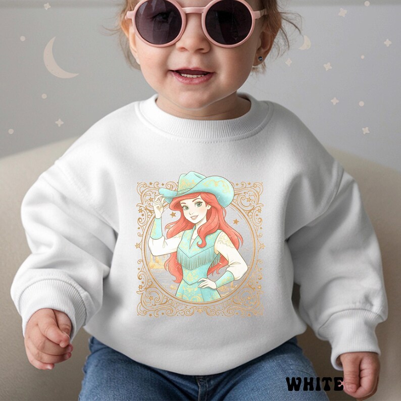 K&ouml;nnte beinhalten: Wei&szlig;es Sweatshirt mit einer Cartoon-Illustration einer Frau mit Cowboyhut und t&uuml;rkisfarbener Weste. Die Illustration ist in einem goldenen, verzierten Design gerahmt. Das Wort "WHITE" ist unten aufgedruckt.