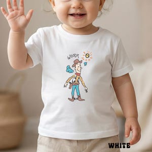 May include: White t-shirt featuring a cartoon illustration of Woody from Toy Story. The design includes Woody in a cowboy hat, a yellow plaid shirt, blue pants, and brown boots. The word "WOODY" is written above the character.