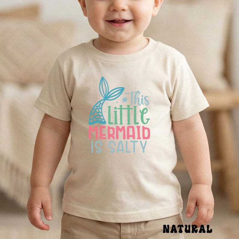 Puede incluir: Camiseta beige para ni&ntilde;o peque&ntilde;o con el texto "This Little Mermaid Is Salty" en turquesa, verde, rosa y azul. La camiseta presenta un gr&aacute;fico de cola de sirena. La palabra "NATURAL" est&aacute; impresa en la parte inferior de la camiseta.