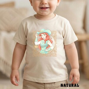 K&ouml;nnte beinhalten: Beiges T-Shirt mit einer Cartoon-Illustration einer Cowgirl mit roten Haaren, t&uuml;rkisfarbenem Hut und Weste, in einem goldfarbenen Rahmen. Das Wort "NATURAL" ist unten in Schwarz gedruckt. Kinderbekleidung.