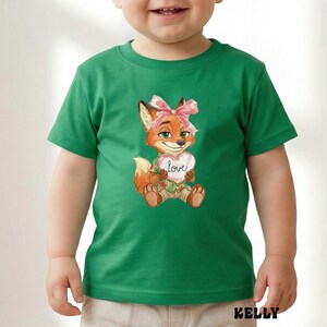 Peut inclure: T-shirt vert avec une illustration &agrave; l'aquarelle d'un renard tenant un c&oelig;ur avec le mot "Love". Le renard porte un n&oelig;ud rose et une chemise &agrave; fleurs. Le mot "KELLY" est imprim&eacute; en bas.