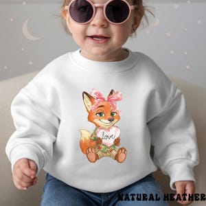Peut inclure: Sweat-shirt blanc avec un renard de dessin anim&eacute; tenant un c&oelig;ur avec le mot "love". Le renard porte un n&oelig;ud rose et une chemise hawa&iuml;enne. Le texte "NATURAL HEATHER" est en bas.