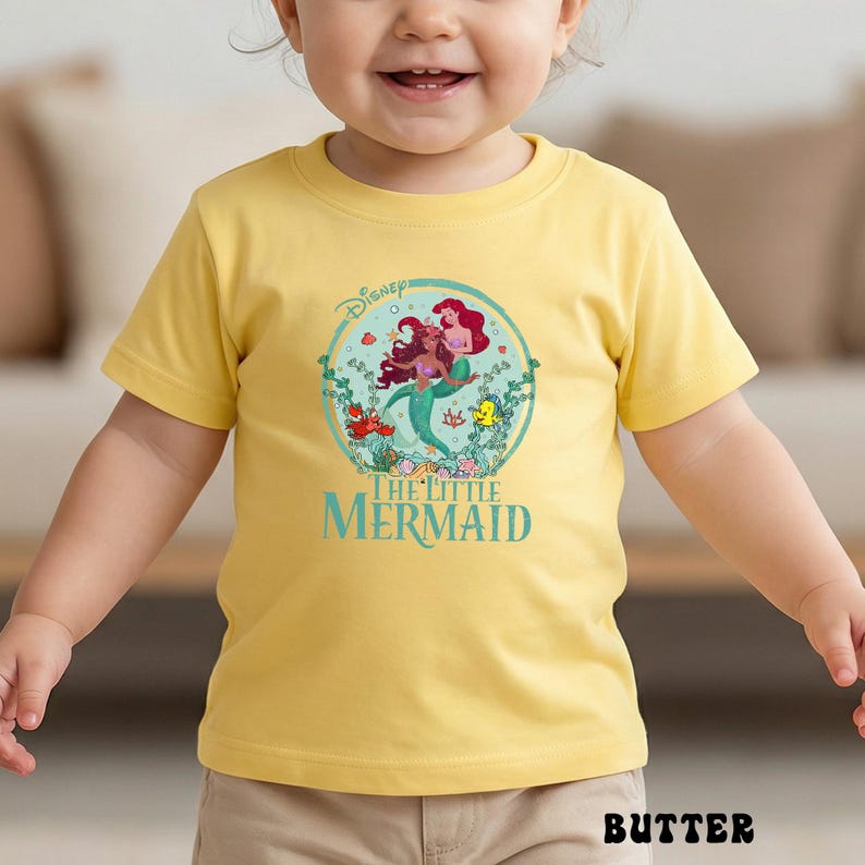May include: A yellow t-shirt featuring a graphic of Ariel and another mermaid from Disney's The Little Mermaid. The design is in a circular frame with the words "The Little Mermaid" below. The shirt is worn by a child.