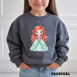 Puede incluir: Una sudadera gris carb&oacute;n de cuello redondo con un gr&aacute;fico central de un personaje de dibujos animados con largo cabello rojo, corpi&ntilde;o morado y vestido de gala verde claro. La sudadera tiene mangas largas con pu&ntilde;os y cuello acanalados.