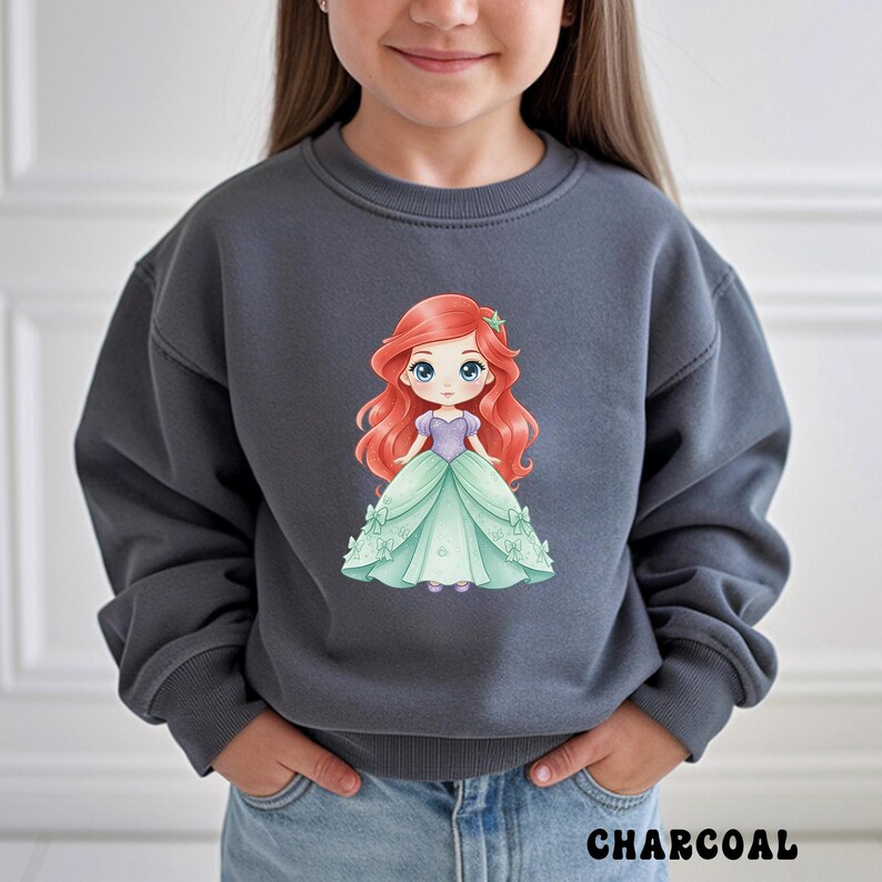 Puede incluir: Una sudadera gris carb&oacute;n de cuello redondo con un gr&aacute;fico central de un personaje de dibujos animados con largo cabello rojo, corpi&ntilde;o morado y vestido de gala verde claro. La sudadera tiene mangas largas con pu&ntilde;os y cuello acanalados.
