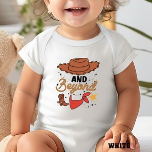 May include: White baby bodysuit with a cowboy-themed graphic. The design includes a brown cowboy hat, the words "AND Beyond" in a decorative font, a cowboy boot, a red bandana, and a yellow star. The word "WHITE" is printed at the bottom.