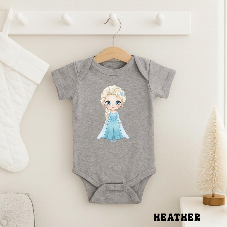 Puede incluir: Un body de beb&eacute; gris con una ilustraci&oacute;n de dibujos animados de una princesa con un vestido azul. La princesa tiene el pelo rubio y ojos azules. La palabra "HEATHER" est&aacute; impresa en negro en la parte inferior de la imagen.