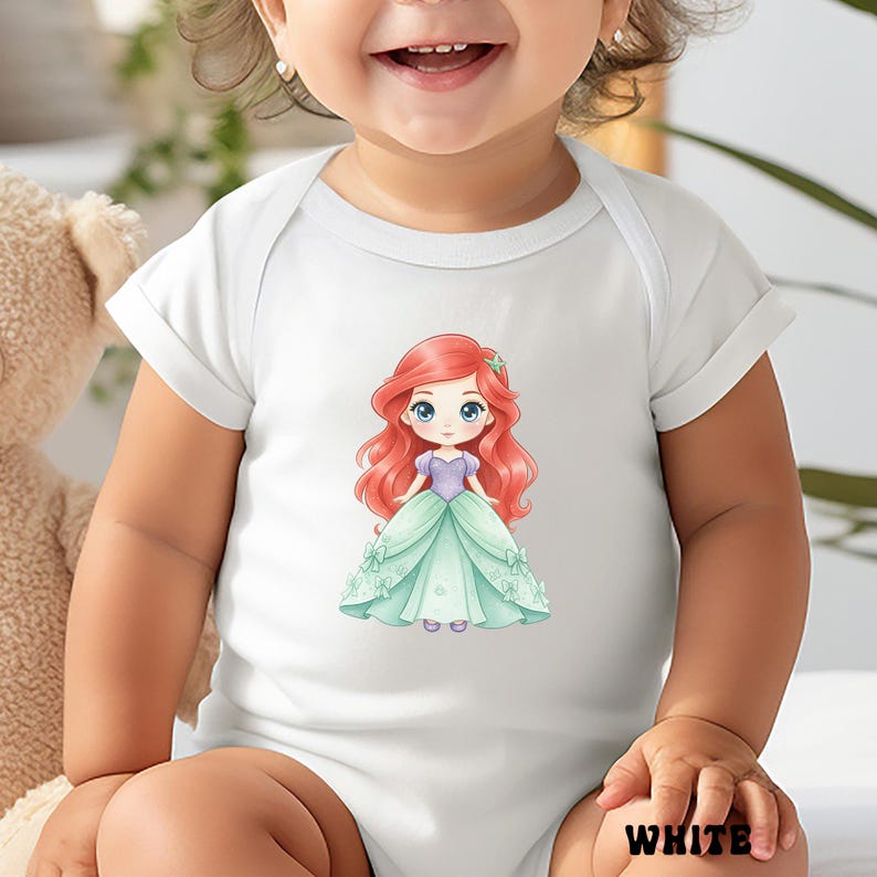 Puede incluir: Un body blanco de manga corta para beb&eacute; con un gr&aacute;fico central de un personaje de dibujos animados con largo cabello rojo, corpi&ntilde;o morado y vestido verde claro. El personaje tiene grandes ojos azules y un peque&ntilde;o accesorio de estrella verde.