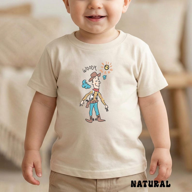 May include: A beige t-shirt featuring a cartoon illustration of Woody from Toy Story. The design includes Woody in a cowboy hat, blue pants, and a red neckerchief. The word "WOODY" is written above the character. The word "NATURAL" is printed at the bottom.
