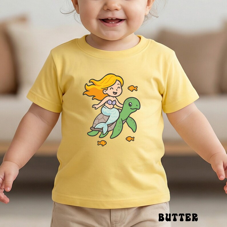 Puede incluir: Camiseta amarilla para ni&ntilde;os peque&ntilde;os con una sirena de dibujos animados montada en una tortuga verde, rodeada de peces naranjas. La sirena tiene el pelo rubio y una cola azul. La palabra "BUTTER" est&aacute; impresa en la parte inferior.