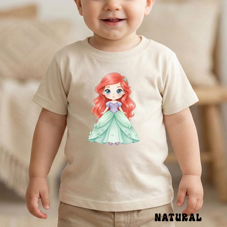 Puede incluir: Una camiseta beige de manga corta con una ilustraci&oacute;n gr&aacute;fica centrada de un personaje de dibujos animados con largo cabello rojo, que viste un corpi&ntilde;o morado y un vestido de gala verde claro con peque&ntilde;os detalles de lazos.