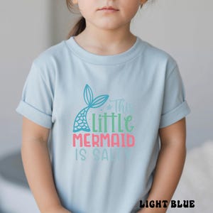Puede incluir: Camiseta azul claro con mangas enrolladas. La camiseta presenta un dise&ntilde;o de cola de sirena y el texto "This Little Mermaid Is Salty" en verde azulado, verde y rosa. Las palabras "LIGHT BLUE" est&aacute;n en la parte inferior de la camiseta.
