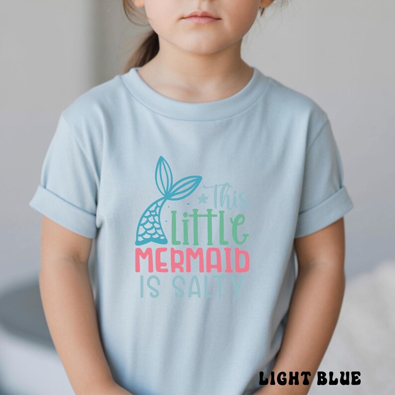 Puede incluir: Camiseta azul claro con mangas enrolladas. La camiseta presenta un dise&ntilde;o de cola de sirena y el texto "This Little Mermaid Is Salty" en verde azulado, verde y rosa. Las palabras "LIGHT BLUE" est&aacute;n en la parte inferior de la camiseta.