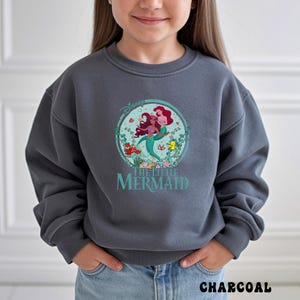 May include: Charcoal gray sweatshirt featuring a circular graphic of Ariel and another mermaid from Disney's The Little Mermaid. The graphic includes the Disney logo and the words "THE LITTLE MERMAID" in teal. The word "CHARCOAL" is printed at the bottom.