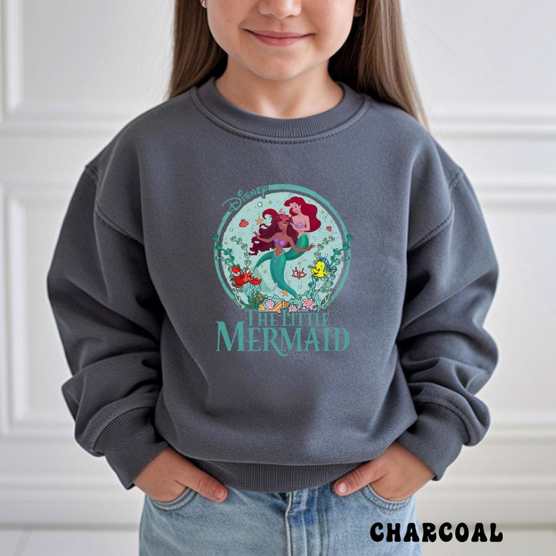 May include: Charcoal gray sweatshirt featuring a circular graphic of Ariel and another mermaid from Disney's The Little Mermaid. The graphic includes the Disney logo and the words "THE LITTLE MERMAID" in teal. The word "CHARCOAL" is printed at the bottom.