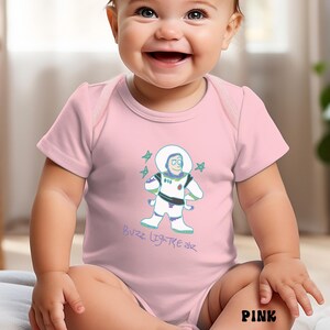 May include: Pink baby bodysuit featuring a Buzz Lightyear graphic. The illustration shows Buzz in his space suit with stars. The text "Buzz Lightyear" is written below the graphic. The word "PINK" is printed at the bottom.