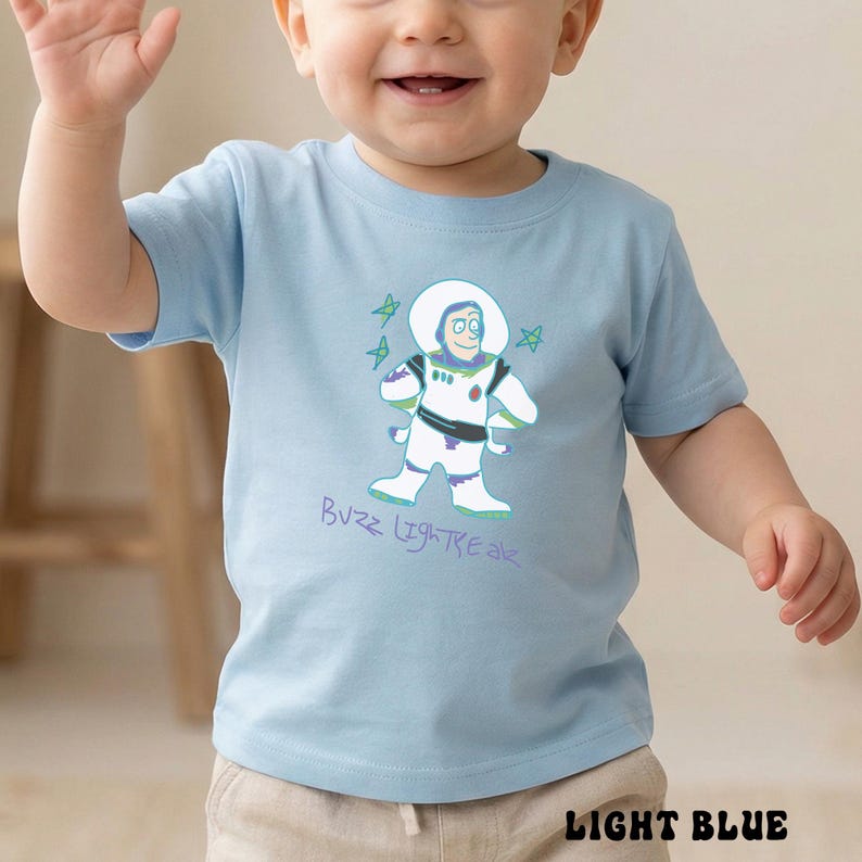 May include: Light blue toddler t-shirt featuring a cartoon illustration of Buzz Lightyear with the text "Buzz Lightyear". The design includes a white spacesuit, green accents, and stars. The shirt is light blue.