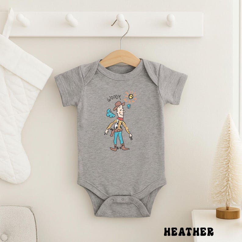 May include: A heather gray baby onesie featuring a cartoon illustration of Woody from Toy Story. The design includes Woody's name, hat, and outfit. The onesie is hanging on a wooden hanger.