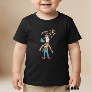 May include: Black t-shirt featuring a cartoon illustration of Woody from Toy Story. The design includes the word "WOODY" above the character, along with a sun and butterfly graphic. The word "BLACK" is printed at the bottom of the shirt.