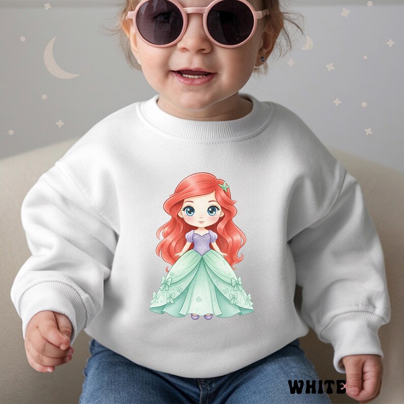 Puede incluir: Una sudadera blanca de manga larga con un gr&aacute;fico central de un personaje de dibujos animados con largo cabello rojo, un corpi&ntilde;o morado y un vestido verde claro. El personaje tiene grandes ojos azules y un peque&ntilde;o accesorio de estrella.