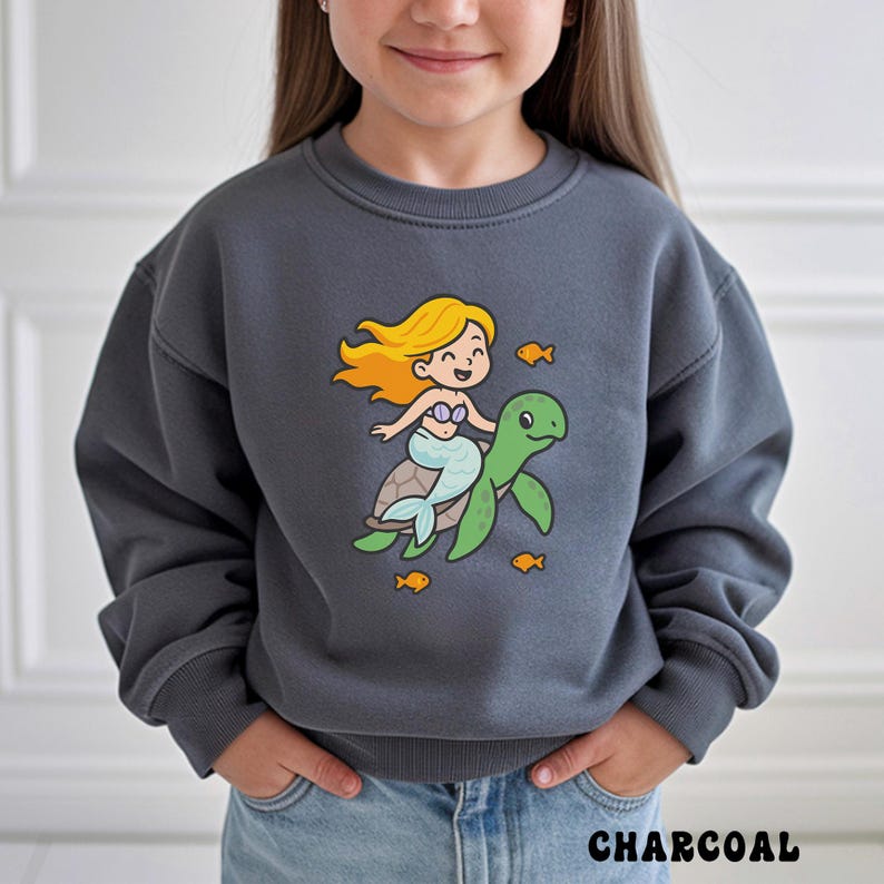 Puede incluir: Sudadera gris carb&oacute;n con una sirena de dibujos animados montada en una tortuga verde, rodeada de peces naranjas. La sirena tiene el pelo rubio y una cola azul claro. La palabra "CHARCOAL" est&aacute; impresa en la parte inferior de la imagen.
