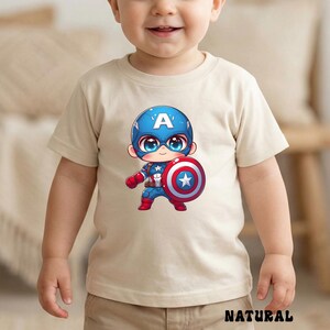 Op de afbeelding: Een beige t-shirt met een cartoon Captain America-afbeelding. De illustratie toont Captain America in een blauwe helm met een witte "A", die zijn schild vasthoudt. Het woord "NATURAL" staat onderaan het shirt.