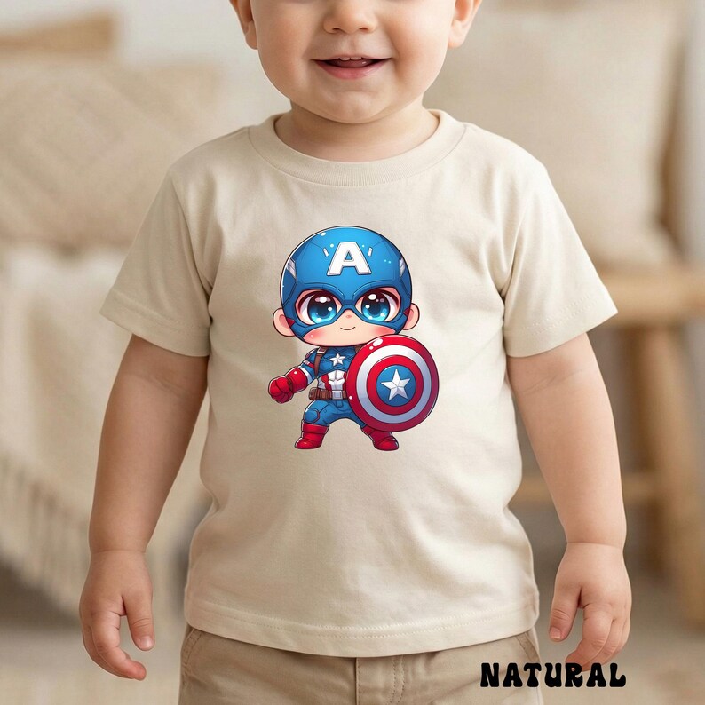 Op de afbeelding: Een beige t-shirt met een cartoon Captain America-afbeelding. De illustratie toont Captain America in een blauwe helm met een witte "A", die zijn schild vasthoudt. Het woord "NATURAL" staat onderaan het shirt.