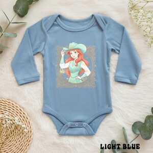 K&ouml;nnte beinhalten: Hellblaues Baby-Body mit langen &Auml;rmeln. Auf der Vorderseite befindet sich eine gerahmte Illustration einer Cowgirl mit roten Haaren und einem t&uuml;rkisfarbenen Hut und Outfit. Der Text "LIGHT BLUE" ist unten aufgedruckt.
