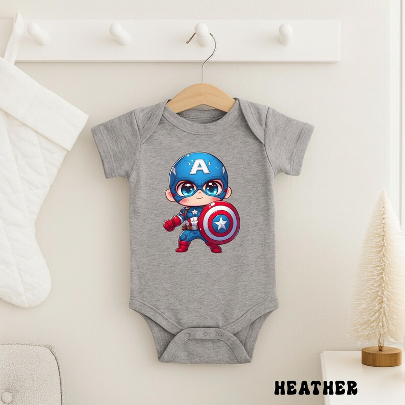 Op de afbeelding: Een grijze babybody met een Captain America cartoonontwerp. De illustratie toont Captain America in een blauwe helm met een witte "A", die een schild vasthoudt. Het woord "HEATHER" staat onderaan afgedrukt.