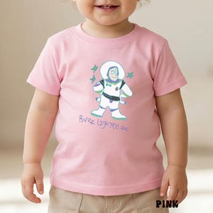 May include: Pink short-sleeved t-shirt featuring a cartoon illustration of Buzz Lightyear with the text "Buzz Lightyear" in a light blue color. The design includes stars and the word "PINK" at the bottom.