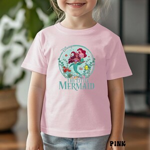 May include: Pink t-shirt featuring a circular graphic of Ariel and another mermaid from Disney's The Little Mermaid. The graphic includes the Disney logo and the words "The Little Mermaid" in teal. The shirt is short-sleeved.