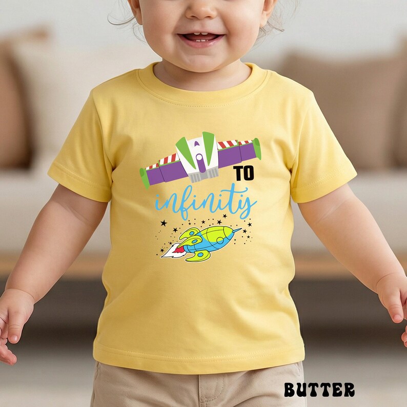 May include: A yellow t-shirt with a colorful graphic featuring a spaceship and the words "To infinity" in blue script. The design includes a spaceship and stars. The word "BUTTER" is printed at the bottom.
