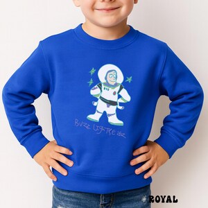 May include: Royal blue sweatshirt featuring a cartoon illustration of Buzz Lightyear in a spacesuit. The design includes the text "Buzz Lightyear" in a handwritten style. The sweatshirt is a solid royal blue color.