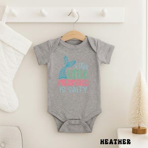 Puede incluir: Un body de beb&eacute; gris jaspeado con un gr&aacute;fico de cola de sirena en turquesa y un texto rosa y turquesa que dice "This Little Mermaid Is Salty". El body est&aacute; colgado de una percha de madera.
