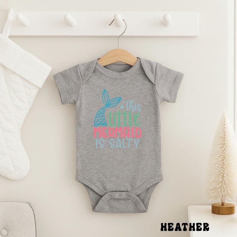 Puede incluir: Un body de beb&eacute; gris jaspeado con un gr&aacute;fico de cola de sirena en turquesa y un texto rosa y turquesa que dice "This Little Mermaid Is Salty". El body est&aacute; colgado de una percha de madera.