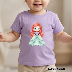 Puede incluir: Una camiseta de manga corta de color morado claro con un gr&aacute;fico central de un personaje de dibujos animados con el pelo largo y rojo, que lleva un corpi&ntilde;o morado y un vestido de gala verde claro con peque&ntilde;os lazos. La palabra LAVENDER est&aacute; en negro.