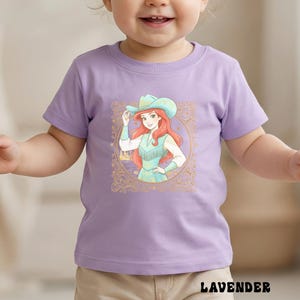 K&ouml;nnte beinhalten: Ein lilafarbenes T-Shirt mit einer Grafik einer rothaarigen Figur mit t&uuml;rkisfarbenem Cowboyhut und Kleid, umrahmt von einem goldenen, verzierten Rand. Das Wort "LAVENDER" ist unten auf dem Shirt aufgedruckt.